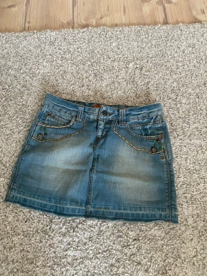 Blå jeanskjol med dekorativa knappt - Säljer en skitsnygg kort jeanskjol med slitna detaljer och dekorativa knappar vid fickorna. Kjolen har klassiska fickor fram och bak, bälteshällor och kontrastsömmar. Finns inte att köpa helt ny och Storleken är 34, pris kan disskuteras💗💗