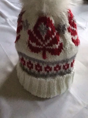 Vit stickad mössa med rött mönster - Stickad mössa i vitt med röda och grå detaljer, dekorerad med ett stort mönster och en fluffig vit boll på toppen. Mössan har bred uppvikt kant och är gjord i mjukt garn, perfekt för kalla dagar. Klassisk vinterstil med retrovibbar.