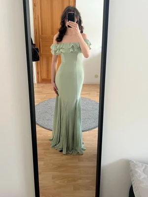 Ljusgrön offshoulder långklänning - Ljusgrön långklänning med offshoulder och volangdetalj upptill. Klänningen har en figurnära passform upptill och faller mjukt ner mot golvet. Jag är 174 cm och har inga skor på bilden. Den är inte använd någon gång.