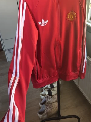 Röd Manchester United Adidas jacka - Säljer en röd träningsjacka från Adidas med Manchester United-logga på bröstet och vit Adidas-logo. Jackan har vita ränder längs ärmarna och texten 'Manchester United' stort på ryggen. Dragkedja framtill och två fickor. Perfekt för fotbollsfans.