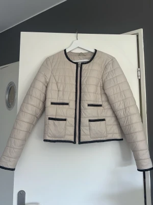 Beige quiltad jacka - Snygg beige quiltad cardiganjacka med svarta detaljer längs kanterna och fickorna. Jackan har rund halsringning, fyra fickor framtill och raka ärmar. Materialet är glansigt och lätt vadderat, vilket ger en modern look. Mkt liten i storlek skulle säga S eller xs och inte medium som lappen visar. Obs säljer endast via plicks köpknapp. 