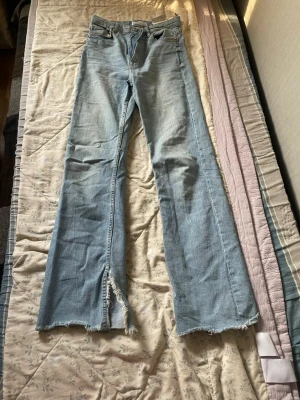 Ljusblå bootcut jeans med slits - Säljer ett par ljusblå bootcut jeans med råa, fransiga benslut och slits framtill. Jeansen har klassisk femficksdesign och hög midja. Perfekta för en avslappnad och trendig look. Materialet är denim i bomull.