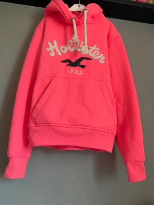 Rosa hoodie från Hollister med huva - Snygg rosa hoodie från Hollister med stor magficka, broderad logga och text på bröstet samt vita dragsnören i huvan. Tillverkad i mjukt material som känns skönt mot huden. Perfekt för en avslappnad och trendig look.