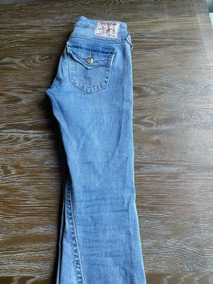 Ljusblå jeans från True Religion - Säljer ett par klassiska ljusblå jeans från True Religion med ikonisk bakficka och snygga detaljer i silver. Jeansen är low wasist bootcut och är tillverkade i mjukt denimtyg som sitter skönt hela dagen.