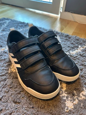 Svarta Adidas sneakers med kardborre - Säljer ett par svarta Adidas sneakers med vita detaljer och tre klassiska ränder på sidan. Skorna har tre kardborreband för enkel påtagning och en vit sula. Tillverkade i syntetmaterial och passar perfekt till en sportig stil.