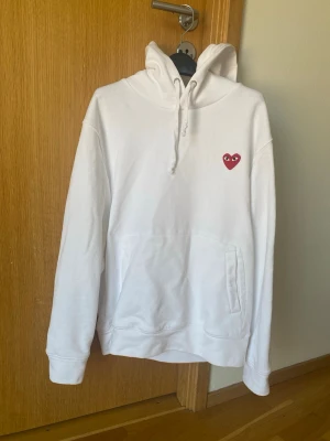 Vit hoodie med rött hjärta - Säljer en vit hoodie med huva och dragsko samt en ficka framtill. På bröstet finns ett litet rött hjärta med ögon som detalj. Hoodien har ribbade muddar vid ärmslut och nederkant. Perfekt för dig som gillar enkel men snygg streetwear.