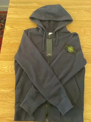 Mörkblå hoodie med dragkedja - Snygg mörkblå hoodie med dragkedja och huva. Har en patch med gul och grön logga på vänster ärm. Utrustad med två fickor framtill och ribbade muddar vid ärmslut och nederkant. Perfekt för en avslappnad streetwear-look.