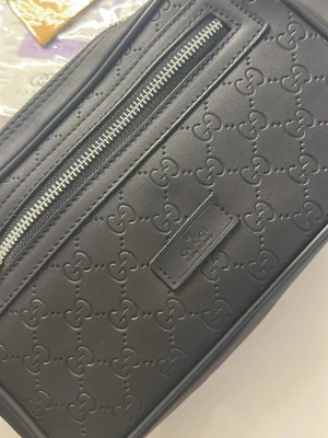 Svart axelväska från Gucci - Snygg svart axelväska från Gucci i läder med GG-mönster över hela väskan. Framsidan har en dragkedja och en diskret logotyp. Baksidan är i mesh och axelbandet är blått med röd rand. Perfekt för dig som vill ha en stilren och lyxig look.