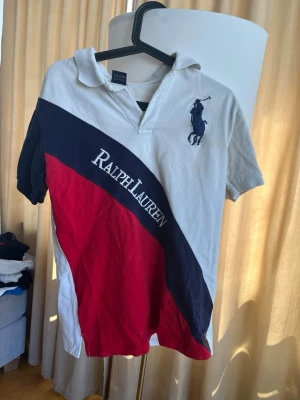 Ralph Lauren vit och röd piké  - Säljer en klassisk skjorta från Ralph Lauren med vit bas, mörkblå diagonal panel och röd detalj. Stort broderat logotyp på bröstet och texten 'Ralph Lauren' över bröstet. Skjortan har korta ärmar och krage, perfekt för en sportig look.