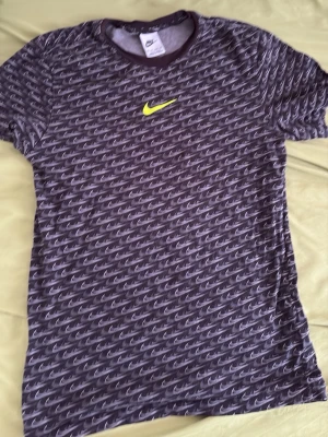 Svart Nike t-shirt med mönster - Snygg svart t-shirt från Nike med all-over print av små vita swoosh-loggor och en större gul swoosh på bröstet. T-shirten har rund hals och korta ärmar. Perfekt för dig som gillar sportig och trendig stil.
