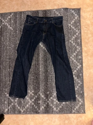 Mörkblå raka jeans John Pearson - Säljer ett par mörkblå jeans från John Pearson & Son med raka ben och klassisk femficksmodell. Jeansen har kontrastsömmar och en snygg läderpatch bak i midjan. Perfekta för dig som gillar tidlös denimstil.