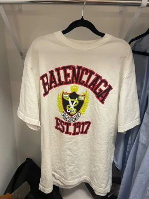 Vit Balenciaga t-shirt med tryck - Vit t-shirt från Balenciaga med stort tryck framtill i rött, svart och gult. Trycket har texten 'Balenciaga EST. 1917' och ett emblem med detaljer. T-shirten har rund hals och korta ärmar, perfekt för en trendig streetwear-look. Inga defekter. Overzized