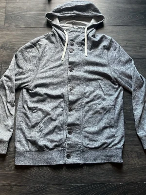 Grå hoodie med knappar från H&M - H&M Hoodie med knappar – Grå – Storlek M  Säljer en stilren H&M-hoodie i grå färg med knäppning framtill och huva. Tröjan är i 9/10 skick, mycket väl omhändertagen och använd sparsamt.  Detaljer:  * Märke: H&M * Storlek: M * Färg: Grå * Skick: 9/10 * Knappar framtill och justerbar huva * Mjuk och bekväm – perfekt för vardag och svalare dagar  Skickas inom 24 timmar. Hör gärna av dig om du har frågor eller vill ha fler bilder!