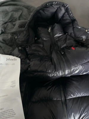Svart dunjacka från ralph lauren äkta - Snygg svart dunjacka med glansig finish och huva från ralph lauren som är äkta jag har använt jackan en fjärdedel av en vinter anledning till att jag säljer den är för att den är för stor för mig jag är (159).             Jackan är äkta och jag har kvar ettiketterna och påsen från johnells där jag köpte den ifrån även kvittot är kvar.