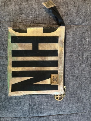 Guldig kuvertväska från Moschino - Snygg kuvertväska från Moschino i guldigt skinn med stora svarta bokstäver på framsidan. Väskan har dragkedja upptill och en metallbricka med Moschino-logga. Perfekt accessoar för att sticka ut.