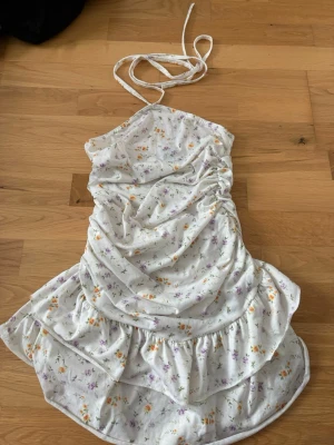 Vit blommig klänning - Söt vit klänning med blommigt mönster i lila och orange. Klänningen har omlottdesign och volangdetaljer nertill. Perfekt för varma dagar och ger en romantisk känsla. Inte använd utan bara prövat den på 