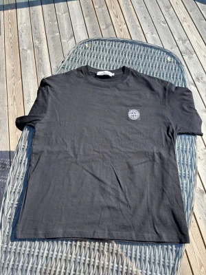 Svart Stone Island t-shirt - Säljer en svart t-shirt från Stone Island med klassisk logopatch på bröstet. T-shirten har rund hals och korta ärmar, tillverkad i mjuk bomull för en skön och avslappnad stil.