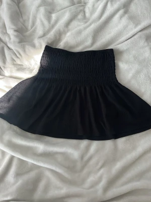 Svart kortkjol från Gina Tricot - Säljer en svart kortkjol från Gina Tricot med smockad midja och lätt utställd passform. Kjolen är gjord i ett mjukt, stretchigt material och har en enkel, stilren look som passar till många olika outfits.