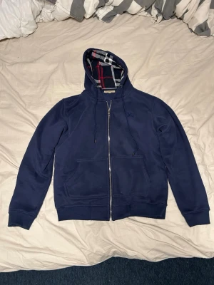 Marinblå hoodie från Burberry - Säljer en marinblå hoodie från Burberry med dragkedja och klassiskt rutigt foder i huvan. Hoodien har två fickor framtill, justerbar dragsko i huvan och mjukt material på insidan. Perfekt för dig som gillar stilrena detaljer och exklusiv känsla.