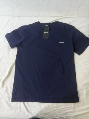Mörkblå t-shirt från BOSS - Snygg mörkblå t-shirt från BOSS med diskret logga på bröstet. Klassisk rund hals och korta ärmar. Tillverkad i mjuk bomull som känns skön mot huden. Perfekt till jeans eller shorts för en clean och stilren look.