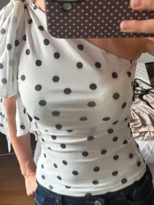 Vit prickig oneshoulder topp - Snygg vit topp med svarta prickar i oneshoulder-modell. Toppen har en stor rosett på axeln och är figurnära med stretchigt material. Perfekt för dig som vill sticka ut med en trendig och lekfull look.