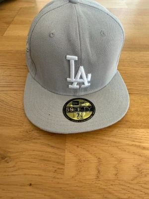 Ljusgrå LA 59FIFTY keps - Snygg ljusgrå 59FIFTY keps från New Era med LA-logga broderad framtill och World Series-detalj på sidan. Klassisk rak skärm och strukturerad passform. Perfekt för dig som gillar streetwear och sportig stil.