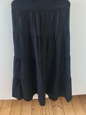 Svart långkjol med volangdetaljer - En svart långkjol med flera volangdetaljer och smock i midjan. Kjolen har en luftig och flowy passform som ger en avslappnad men ändå stilren look. Perfekt att matcha med en enkel topp eller stickad tröja.