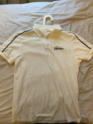 Piké med svarta detaljer XS/S - Vit pikétröja med svarta ränder längs axlarna och broderad text på bröstet. Klassisk krage och korta ärmar. Perfekt för en clean och sportig look.