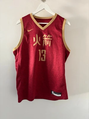 James Harden Chinese New year Linne  - Snygg röd basketlinne med guldiga kanter och tryck, samt nummer 13 på framsidan. Linnet har kinesiska tecken och är ärmlöst med v-ringad hals. Perfekt för dig som gillar sportig stil och vill sticka ut.