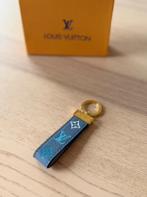 Vuitton nyckelring i blått läder - Säljer en snygg  Vuitton nyckelring i blått och grått läder med klassiska LV-loggor i turkos och vitt. Nyckelringen har guldfärgade metalldetaljer och en rund ring med ingraverad text. Perfekt accessoar för att piffa till dina nycklar eller väska.