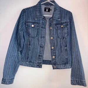 Randig jeansjacka från Boohoo - Snygg blå jeansjacka från Boohoo med vita vertikala ränder. Klassisk modell med krage, bröstfickor med knappar och silverfärgade tryckknappar framtill. Jackan har lång ärm och är tillverkad i jeansmaterial. Perfekt för dig som vill ha en cool och tidlös look.