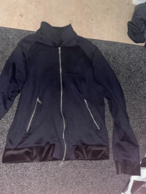 Svart Limitato jacka - Säljer en svart hoodie med dragkedja och två snedställda fickor framtill. På ryggen finns ett stort färgstarkt tryck av ett konstnärligt porträtt i blått och rött. Hoodien har hög krage och muddar i glansigt material vid ärmslut och nederkant.