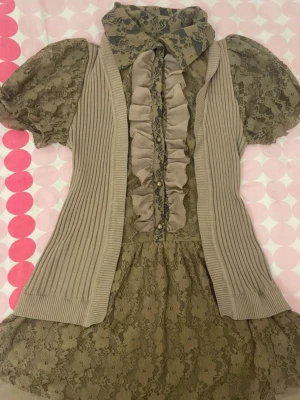 Beige spetsblus med volang och väst - Supersöt beige blus i spets med puffärmar och volangdetalj framtill. Blusen har en ribbad stickad väst i samma ton och en spetskrage som ger en vintagekänsla. Materialet är genombruten spets och mjuk stickad trikå. Perfekt för dig som gillar romantisk och unik stil. Jag har i storlek S och denna passar mig ganska bra