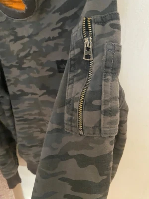 Camo mönstrad sweatshirt med ficka - Säljer en grå och svart camo mönstrad sweatshirt med rund hals och ribbade muddar. Tröjan har en cool ficka med dragkedja på ena ärmen och en liten patch på bröstet. Perfekt för dig som gillar streetwear vibes.