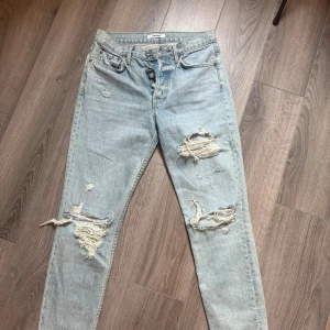 Ljusblå slitna mom jeans - Säljer ett par ljusblå mom jeans med flera stora slitningar och hål på både lår och knän. Jeansen har klassisk femficksmodell, hög midja och raka ben. Perfekta för en avslappnad och trendig look. w26 märke GRFRND
