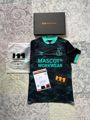 Ny Shamrock Rovers FC fotbollströja - Här har du chansen att köpa en helt ny Shamrock Rovers FC fjärdetröja från säsongen 2024 tillverkad av Umbro med den snygga Mascot Workwear-sponsorn på bröstet. Tröjan levereras i sitt kompletta originalutförande från Mystery United vilket innebär att du får med den eleganta svarta presentationsboxen, det tillhörande informationsbladet om klubben, plastfickan samt alla officiella lappar och taggar. Detta är ett perfekt samlarobjekt eller en fantastisk present för fotbollsälskaren som