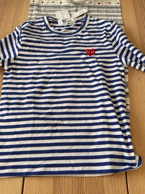 Randig t-shirt med hjärtdetalj - Säljer en blåvit randig t-shirt med korta ärmar och rund hals. På bröstet sitter ett rött broderat hjärta med ögon, vilket ger en unik och lekfull touch. Tillverkad i mjuk bomull som känns skön mot huden. Perfekt för dig som gillar klassiska ränder med en twist.