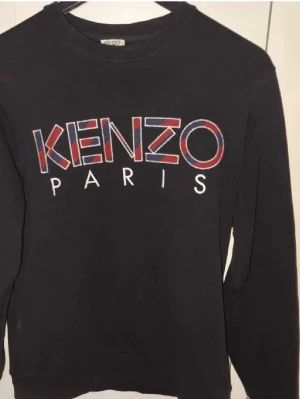 Svart sweatshirt från Kenzo Paris - Svart sweatshirt från Kenzo Paris med stort broderat logotyptryck i rött, blått och vitt på bröstet. Tröjan har rund halsringning och långa ärmar. Tillverkad i mjukt bomullsmaterial som är skönt att bära.