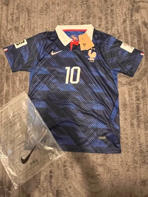 Frankrike Mbappé fotbollströja Nike - Blå fotbollströja från Nike med Frankrikes landslagsemblem, nummer 10 och Mbappé-tryck på ryggen. Tröjan har vit krage med röd detalj, korta ärmar och mönstrad design i olika blå nyanser. Perfekt för dig som älskar fotboll och vill stötta Frankrike.