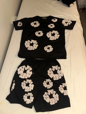 Svart loungewear-set med vita mönster - Säljer ett svart loungewear-set med t-shirt och shorts. Setet har unika vita grafiska mönster i form av cirklar över hela plaggen. Perfekt för chill dagar hemma. Materialet ser ut att vara mjukt och bekvämt, troligen polyester eller bomullsblandning.