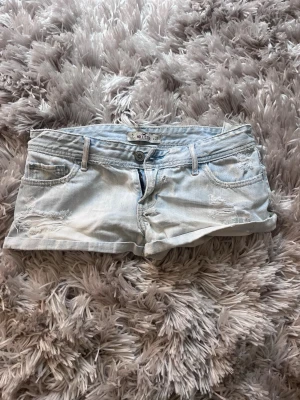 Ljusblå jeansshorts från Hollister - Säljer ett par ljusblå jeansshorts från Hollister med slitna detaljer och klassisk femficksmodell. Shortsen har låg midja, knapp- och dragkedjestängning samt råa kanter nedtill. Perfekta för varma dagar och en avslappnad stil. Har hål men som ser änka ut att laga