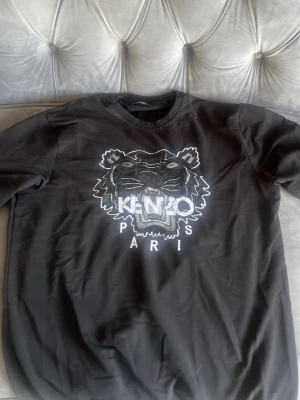 Svart sweatshirt med tigertryck Kenzo - Svart sweatshirt från Kenzo med ikoniskt tigerhuvud broderat framtill och texten 'KENZO PARIS' i vitt. Tröjan har rund halsringning och långa ärmar. Perfekt för dig som gillar streetwear och vill sticka ut med en klassisk logga. Storlek Xl som är runt 4 år gammal och passar perfekt för dig med storlek m-l