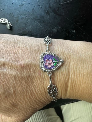 Silverfärgat armband med lila hjärta - Unikt armband i silverfärg med dekorativa blommor längs länken och ett hjärtformat hänge i mitten. Hänget har en lila bakgrund och en rosa blomma med gröna blad. Perfekt för dig som gillar detaljerade och romantiska smycken.