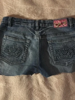 Vintage jeansshorts med detaljer - Snygga vintage lågmidjade jeansshorts med broderade kronor på bakfickorna. De är lite stretchiga och bekväma vilket kan vara väldigt skönt, speciellt när man sitter. Köpta för cirka 20 år sen så de är vintage, unika och går inte att få tag på!! Märkeslapp med rosa detaljer baktill. 