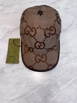 Beige keps med GG-mönster från Gussi - Snygg beige keps från Gussi med det klassiska GG-monogrammet i brunt över hela kepsen. Kepsen har böjd skärm, bruna kanter och justerbar rem bak. Tillverkad i canvasmaterial med insida i ljus textil. Perfekt för dig som gillar exklusiva accessoarer.