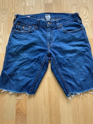 True religon shorts  - Säljer ett par blå jeansshorts med fem fickor och rå, fransig kant nedtill. Shortsen har klassisk knappgylf och kontrastsömmar. Bakfickorna har dekorativa sömmar och lock med knapp. Perfekta för en avslappnad stil.