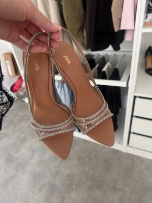 Beige pumps med strass från Zara - Säljer ett par snygga beige pumps från Zara med transparenta remmar dekorerade med strass. Skorna har spetsig tå och smal klack som ger en elegant look. Perfekta för att lyfta vilken outfit som helst.