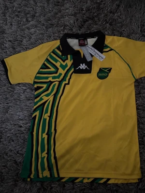 Jamaica fotbollströja från Kappa - Gul kortärmad fotbollströja från Kappa med svart krage och gröna samt svarta grafiska detaljer. Bröstet har Jamaica-märke och Kappa-logga. Tillverkad i lätt och ventilerande material, perfekt för träning eller match.