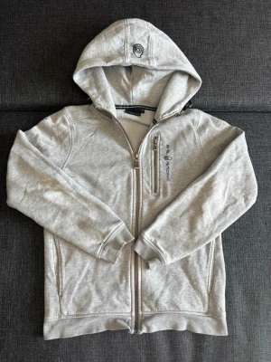 Ljusgrå hoodie från Sail Racing - Ljusgrå hoodie med dragkedja från Sail Racing. Tröjan har huva, två sidofickor och en bröstficka med dragkedja. Broderad logga på bröstet och detaljerade dragkedjor. Perfekt för en avslappnad och sportig stil. Storklek 150