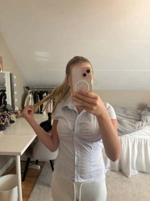 Ljusblå- och vitrandig skjorta - Kortärmad skjorta med ljusblå och vita ränder. Skjortan har klassisk krage, knappar framtill och en figurnära passform. Perfekt för en clean och fräsch look. Man kan även knyta toppen där bak med snörena som är där bak så att det blir till en roset, detta tycker jag själv blir så mycket finare!!